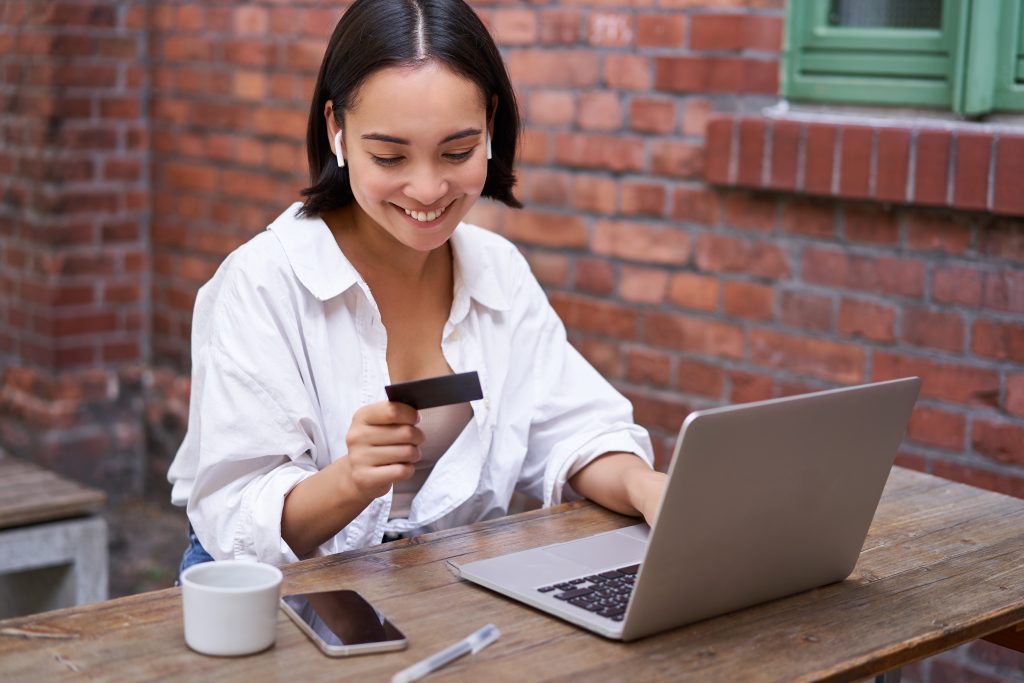 Smiling asian woman with laptop and wireless earphones, paying with credit card, buying online, sitting with cup of coffee