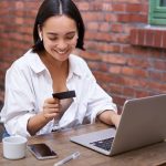 Smiling asian woman with laptop and wireless earphones, paying with credit card, buying online, sitting with cup of coffee